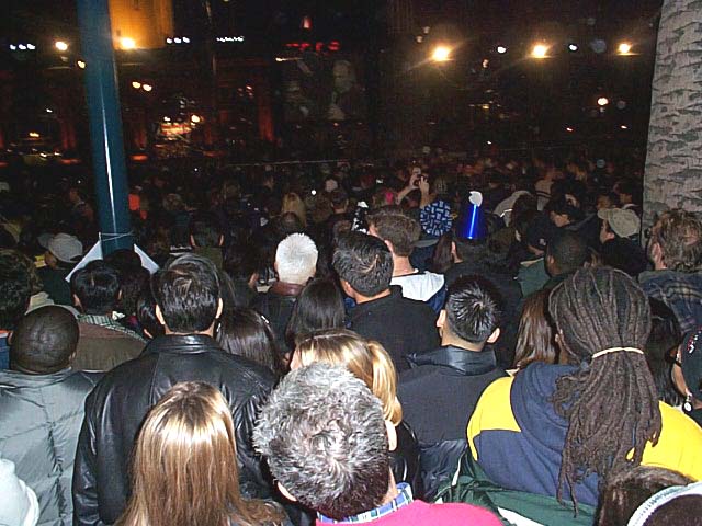 y2k-crowd watches video: The crowd watches a pair of projection-screen TVs, booing at commercials